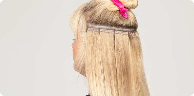 A woman with blonde hair and a pink hair clip showcases her applied tape-in hair extensions.