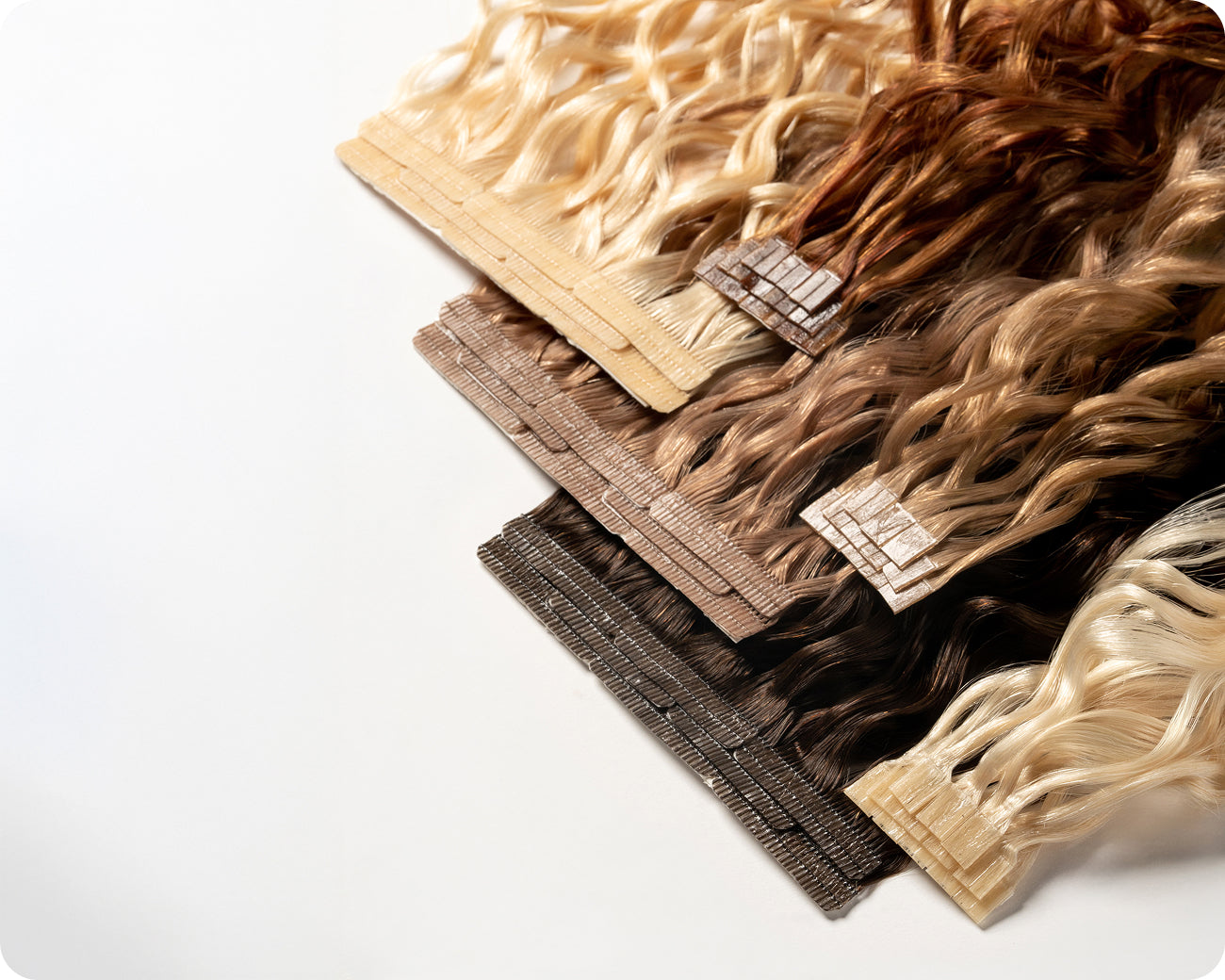 Various shades of wavy tape-in hair extensions are arranged in rows from blonde to dark brown on a white background.
