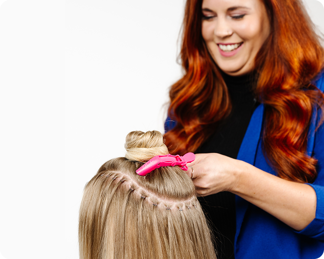 A hairdresser is applying beaded hair extensions to a woman using a pink clip sectioning the hair.