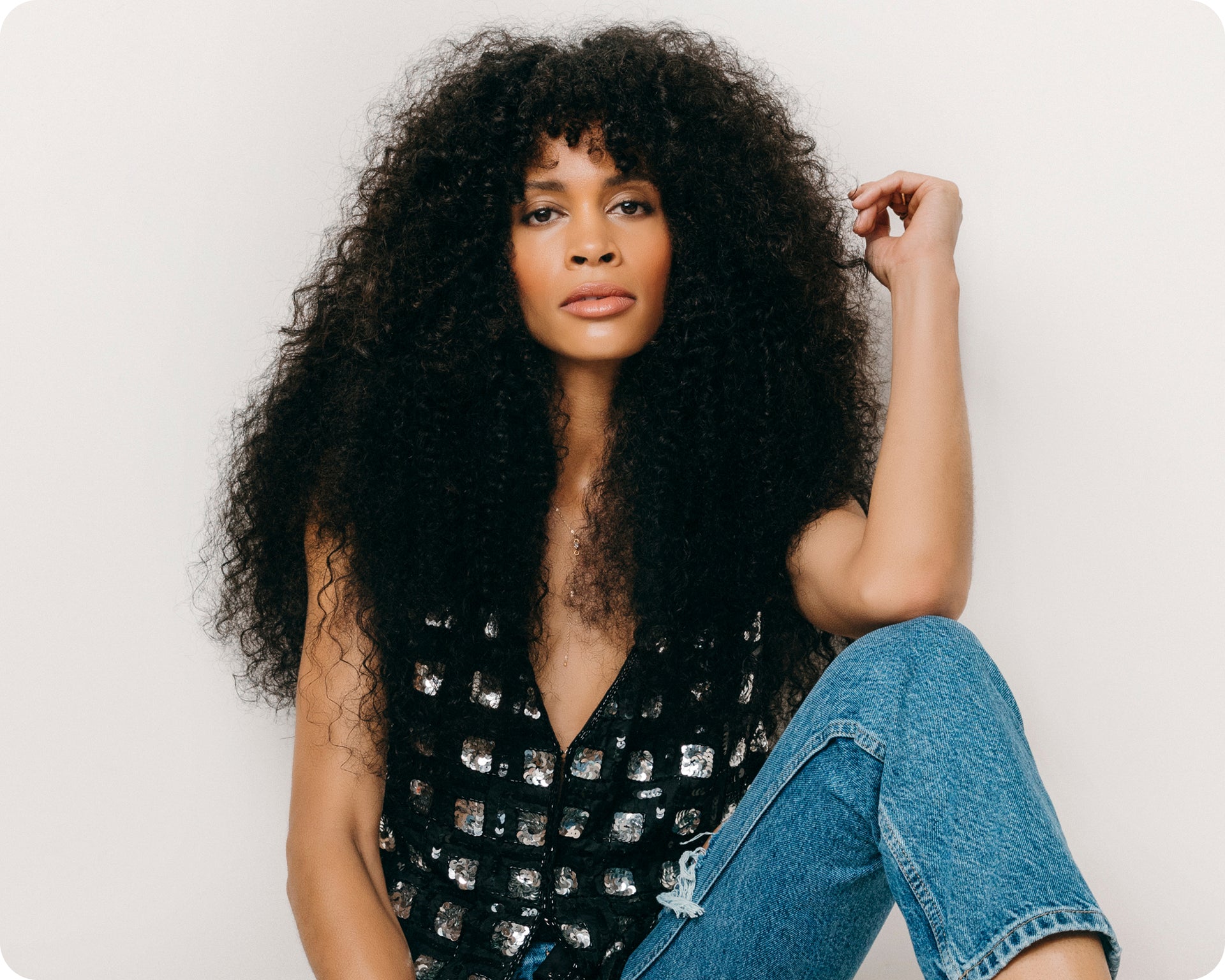 A woman with long, curly, natural black hair extensions sits gracefully against a white background.